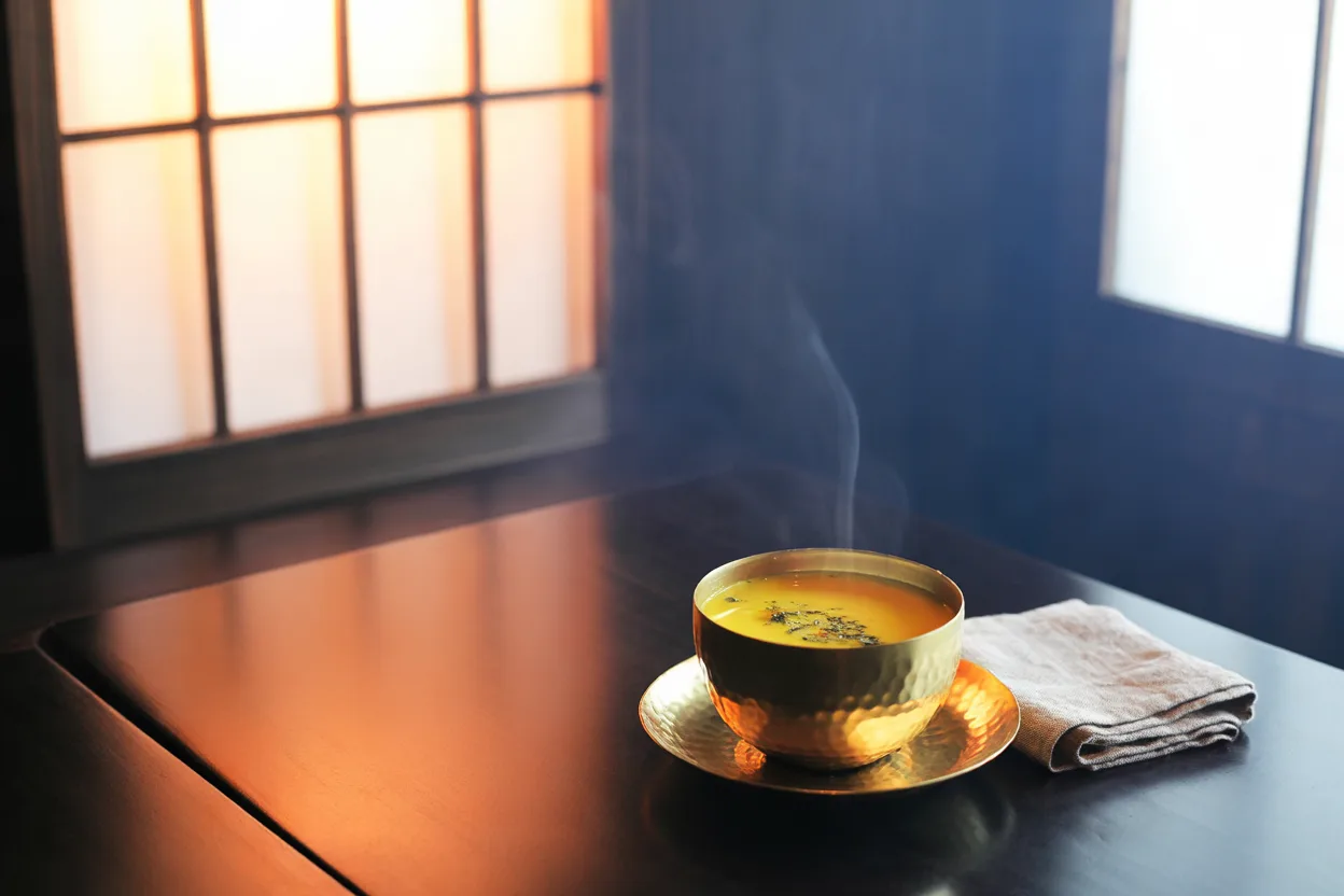 A hammered brass cup of golden milk flecked with black pepper on a dark low table beside a glowing shoji screen at dusk, a folded linen cloth at its side