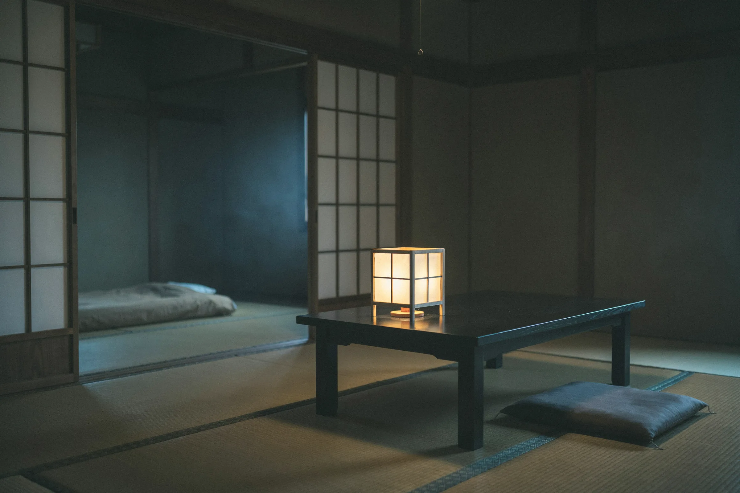 A tatami room at dusk with a paper lantern glowing on a low dark table, an empty cushion beside it and a folded futon in the next room seen through an open shōji screen