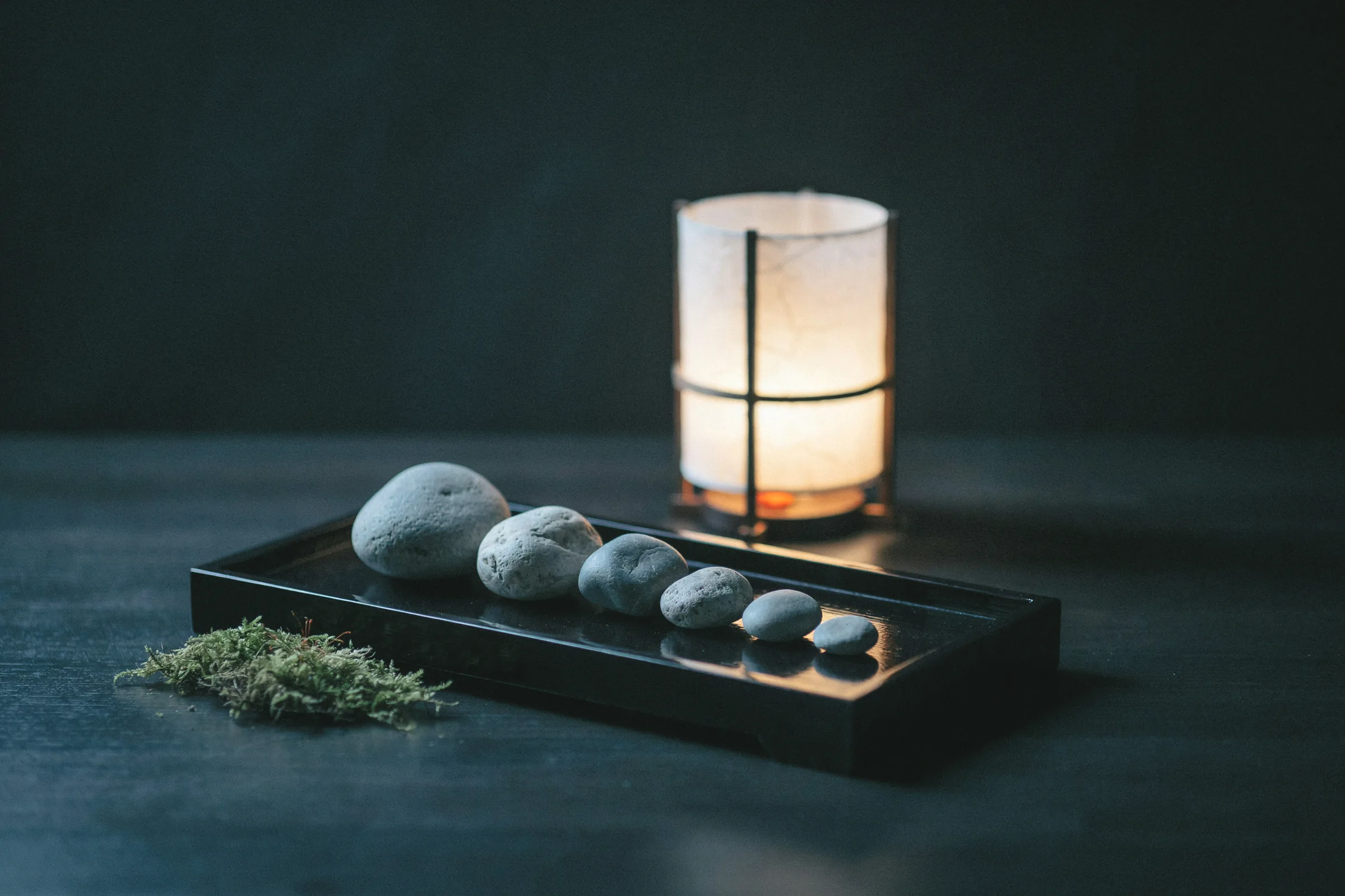 A row of smooth grey river stones shrinking in size along a black lacquer tray, a paper lantern glowing behind and a tuft of moss at the left