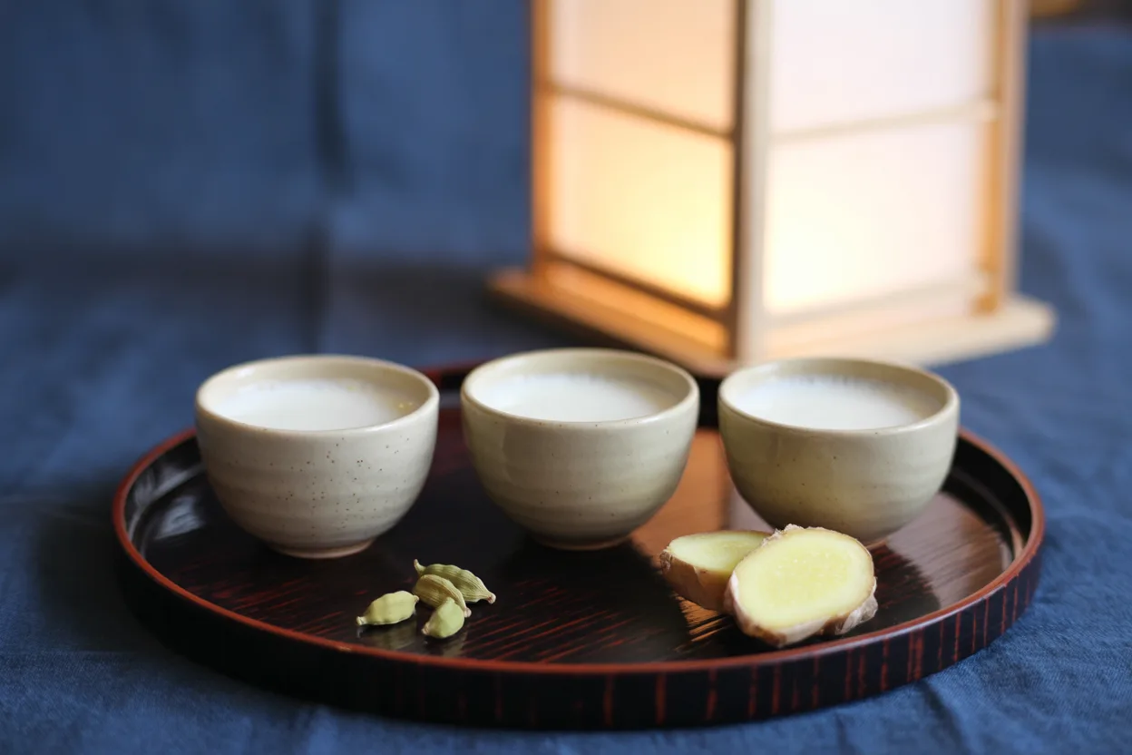 Three small clay cups of warm milk on a round lacquered tray with green cardamom pods and sliced fresh ginger, a paper lantern glowing behind at dusk